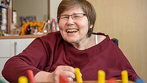 Woman sitting at a table playing a board game. She looks into the camera with a smile.