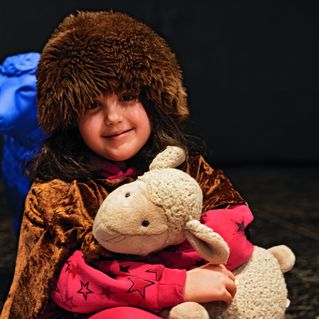 Ein Mädchen hat eine dicke Fellmütze auf dem Kopf, lächelt in die Kamera und hält ein Kuscheltier-Schaf in den Armen