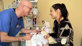 A carer looks after a baby at a hospital bed. The mother stands on the other side of the bed. Both are smiling at each other.