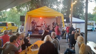 Menschen sitzen an Bierzeltgarnituren und schauen zur Bühne, wo eine Band spielt.