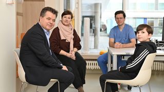 Jürgen Dusel sitzt am Tisch und unterhält sich mit einer Frau, einem Arzt und einem Jungen.