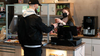 Eine junge Frau steht hinter einer Theke und reicht einem jungen Mann einen Kaffee.