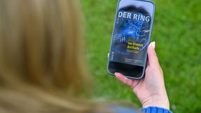 A person is sitting outside holding a smartphone with the digital version of DER RING in her hand.