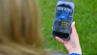 Eine Person sitzt draußen und hält ein Smartphone mit der digitalen Ausgabe von DER RING in den Händen