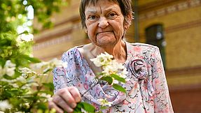 Eine Frau steht an einem  Baum im Garten des Hospizes und hält einen blühenden Strauch in der Hand.