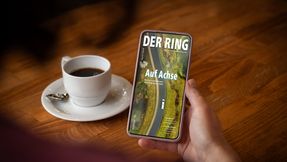 Hand hält ein Smartphone. Auf dem Display ist das Titelbild von DER RING. Im Hintergrund steht eine Tasse Kaffee.