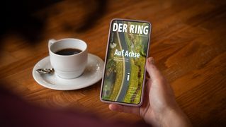 Hand hält ein Smartphone. Auf dem Display ist das Titelbild von DER RING. Im Hintergrund steht eine Tasse mit Kaffee.