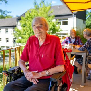 Ein Mann sitzt im Rollstuhl auf einem Balkon und lächelt. 