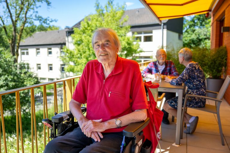 Ein Mann sitzt im Rollstuhl auf einem Balkon und lächelt. 