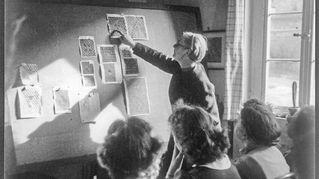 Eine Frau steht vorne an einer Tafel und Schülerinnen hören zu.