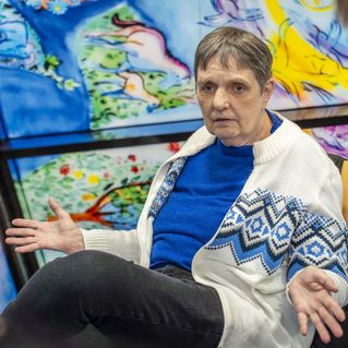 Jutta Ehlers sitzt vor einem Buntglasfenster und gestikuliert. Im Vordergrund sieht man einen jungen Mann, der in ihre Richtung schaut.