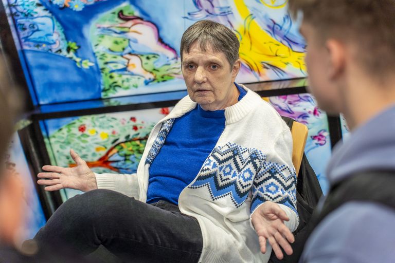 Jutta Ehlers sitzt vor einem Buntglasfenster und gestikuliert. Im Vordergrund sieht man einen jungen Mann, der in ihre Richtung schaut.