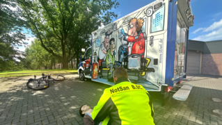 Notfallsanitäter übt Rettungseinsatz an einer Rettungspuppe. Im Hintergrund steht ein Rettungswagen und ein Fahrrad liegt auf der Seite.