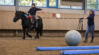 Mädchen reitet auf Pferd und hält sich mit einer Hand fest. Frau hält das Pferd an der Longe.