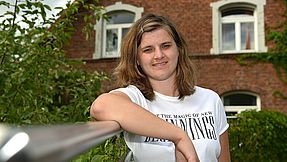 Young woman stands in front of a house and smiles into the camera.