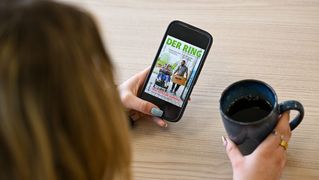 Eine Person sitzt mit einer Tasse Kaffee am Tisch udn liest auf dem Smartphone die digitale Ausgabe von DER RING.