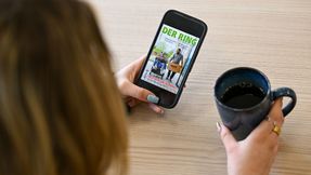 Eine Person sitzt an einem Tisch mit einer Tasse Kaffee und hält ein Smartphone mit der digitalen Ausgabe von DER RING in der Hand.