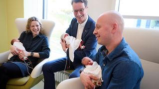 Hendrik Wüst hält ein Baby im Arm, neben ihm sitzen die Mutter und der Vater und halten ebenfalls eins der Drillinge auf dem Arm.