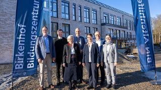 Eine Gruppe Männer und Frauen steht vor dem Neubau. Links und rechst neben der Gruppe stehen Fahnen mit der Aufschrift "Birkenhof Bildungszentrum" und "BIN Altenhilfe".