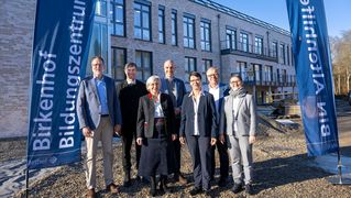 Eine Gruppe Männer und Frauen steht vor dem Neubau. Links und rechst neben der Gruppe stehen Fahnen mit der Aufschrift "Birkenhof Bildungszentrum" und "BIN Altenhilfe".