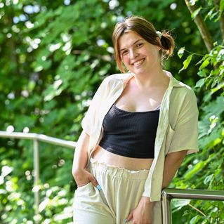 Alicia Lütze steht an einem Geländer vor grünen Büschen und lächelt in die Kamera.