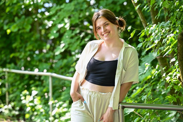 Alicia Lütze steht an einem Geländer vor grünen Büschen und lächelt in die Kamera.