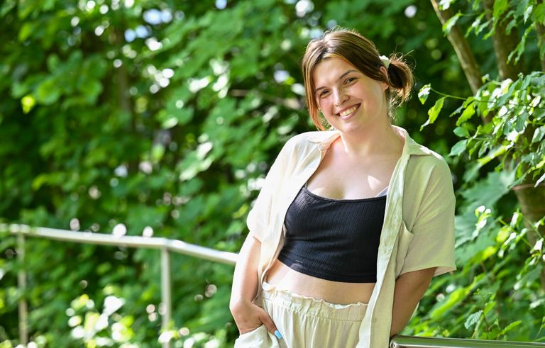 Alicia Lütze steht an einem Geländer vor grünen Büschen und lächelt in die Kamera.