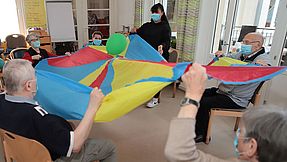 Bewohnerinnen und Bewohner sitzen im Stuhlkreis und halten Luftballon mit einem Schwungtuch hoch.