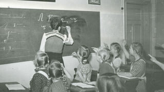 Schwarz-weiß-Foto: eine Lehrerin schreibt an eine Tafel, sieben Mädchen schauen zur Tafel.