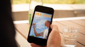 Eine Person sitzt an einem Tisch auf dem ein Glas Wasser steht und hält ein Smartphone mit der digitalen August-Ausgabe von DER RING in der rechten Hand.