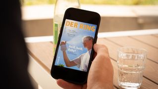 Eine Person sitzt an einem Tisch mit einem Glas Wasser und hält ein Smartphone mit der neuesten Ausgabe von DER RING in der Hand.