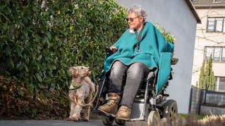 Ursula Niggemeier sitzt im Rollstuhl und führt ihren Hund Gassi zwischen Wohnhäusern und einer Hecke.