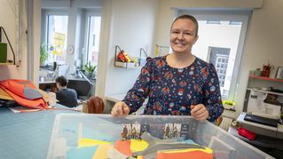 Eine Frau steht an einer Kiste mit Schneidermaterial und lächelt dabei in die Kamera