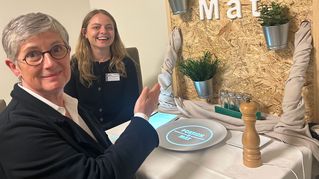 Britta Haßelmann sitzt an einem Tisch und testet unter Anleitung von Anna Katharina Dimas den Portion-o-mat.