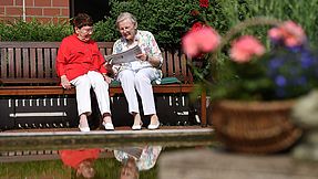 Zwei Seniorinnen sitzen auf einer Bank und lesen Zeitung.