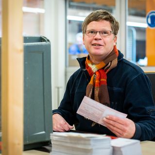 Markus Trommer sitzt an seinem Arbeitsplatz und sortiert Briefe. Freundlich lächelt er in die Kamera