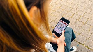 Eine Frau sitzt auf einer Mauer und hält in der Hand ein Smartphone mit dem Cover von DER RING