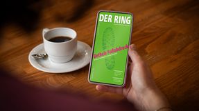 Eine Person sitzt an einem Tisch mit einer Tasse Kaffee und hält ein Smartphone mit der digitalen Ausgabe von DER RING in der Hand.