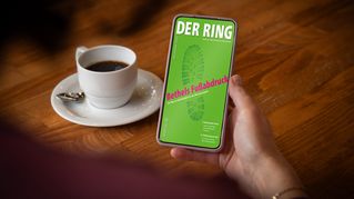 Eine Person sitzt an einem Tisch mit einer Tasse Kaffee und hält ein Smartphone mit der digitalen Ausgabe von DER RING in der Hand.