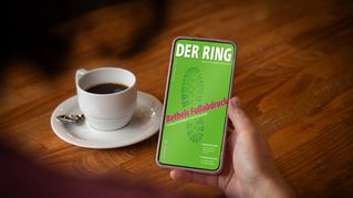 Eine Person sitzt an einem Tisch mit einer Tasse Kaffee und hält ein Smartphone mit der digitalen Ausgabe von DER RING in der Hand.