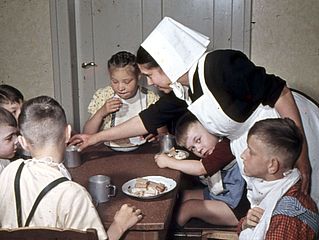 Kinder sitzen am Tisch und eine Diakonisse gibt ihnen Essen. Das Bild entstand in den 1960er-Jahren.