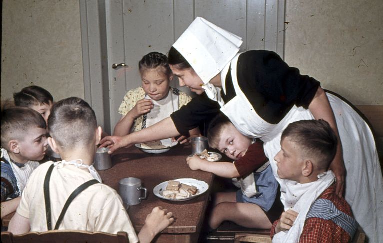 Kinder sitzen am Tisch und eine Diakonisse gibt ihnen Essen. Das Bild entstand in den 1960er-Jahren.