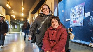 Eine Frau steht hinter einer Frau im Rollstuhl in einer Citypassage. Im Hintergrund sind Filmplakate und leuchten Lichter.