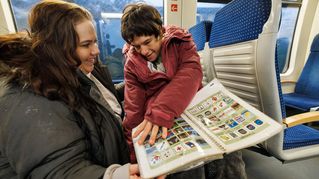 Eine Frau im Rollstuhl blättert in einer Mappe mit vielen Symbolen. Eine andere Frau hat sich zu ihr gehockt.