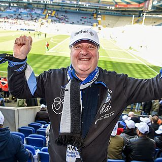 Rainer Wollenburg steht auf der Tribühne im Fußball-Stadion und hält jubelnd beide Arme in die Höhe.