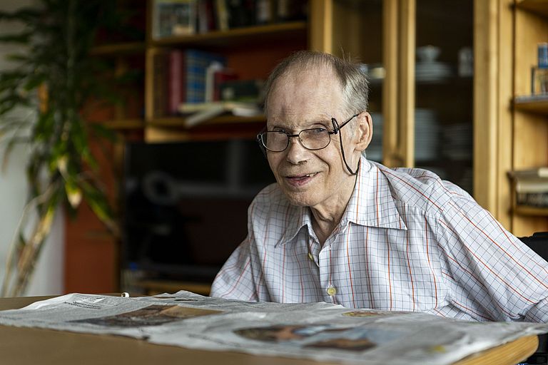 Anton Bals sitzt über eine Zeitung gebeugt am Tisch und lächelt in die Kamera.