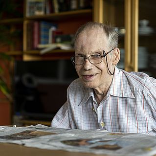 Anton Bals sitzt über eine Zeitung gebeugt am Tisch und lächelt in die Kamera.