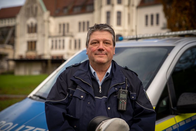 Dirk Hackländer steht in Polizeiuniform vor seinem Polizeiauto. Im Hintergrund sieht man das Haus Sarepta.