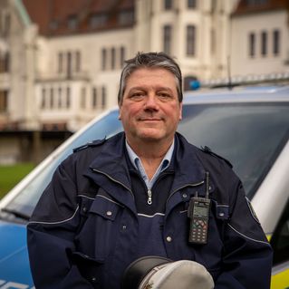 Dirk Hackländer steht in Polizeiuniform vor seinem Polizeiauto. Im Hintergrund sieht man das Haus Sarepta.