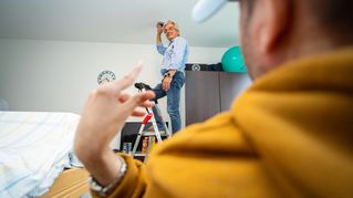 Mann bringt auf Leiter ein Raum-Monitoring-System an die Decke des Klienten an - dieser schaut zu.